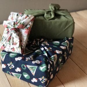Photo de 3 cadeaux emballés dans des Furoshikis, emabllage de cadeaux en tissu, réutilisation à l'infini aux motifs variés : un uni kaki, un aux motifs de noël et coloris blanc et rouge et le dernier aux motifs de Noël dans les ton vert rouge et bleu marine.